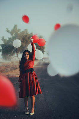 young brown-skinned woman in red dress, black belt, and white shoes releases red and white balloons