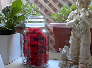 Windowsill looking out to a trellis (left to right) green plant in white pot, glass jar containing crochetd poppies, terracotta-coloured windowbox with geranium foliage, statue of boy with bunnies.