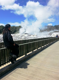 Man sitting on a railing of a bridge, watching tourists taking photos of a geyser.