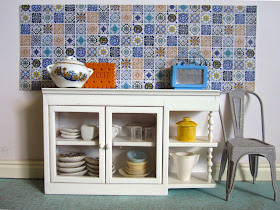 Modern dolls house miniature kitchen scene with a white glass-fronted cupboard full of crockery in front of a wall tiled with various blue and white tiles. Next to teh cupboard is a french cafe chair and on top is a tureen, a giant ornamental biscuit and a Roberts-style radio in blue.