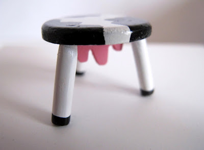 Modern dolls' house miniature stool, painted in cow print, with a pink wooden piece in the shape of an udder underneath the seat.