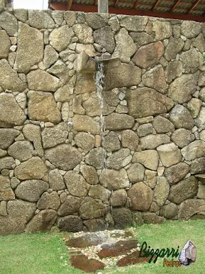 Tipo de muro de pedra com pedra bruta com a ducha d'água na bica de pedra com o piso de pedra em baixo da ducha com os pedregulhos do rio e o gramado de grama esmeralda.