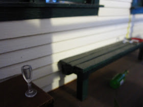 Early-morning shot of a one-twelfth scale model school veranda, with a school chair with an empty wine glass on its seat and a bench with a toppled glass on it and an empty wine bottle under it.
