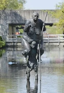 Survival of the Fattest is a sculpture of a small starving African man, carrying Lady Justice, a huge obese European woman who is a symbol of the rich world.