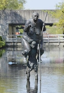 Survival of the Fattest is a sculpture of a small starving African man, carrying Lady Justice, a huge obese European woman who is a symbol of the rich world. Survival of the Fattest is a sculpture of a small starving African man, carrying Lady Justice, a huge obese European woman who is a symbol of the rich world.