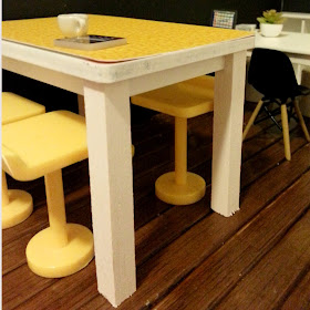 One-twelfth scale modern miniature high work table with white wooden legs and a yellow printed top. Around it are yellow high stools.