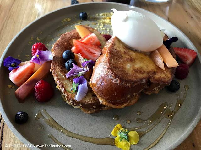 French toast decorated with edible flowers and sliced strawberries and peaches on a grey breakfast plate