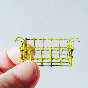 Hand holding up a one-twelfth scale modern miniature wire basket with handles, painted yellow.