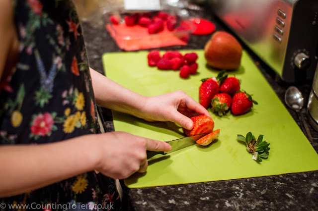 A young girl in a pretty blue flowery dress cutting up a strawberry on a green chopping board