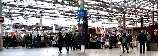 A wide view of the market under the roof of the Main Assembly Building (MAB) at Tonsley. The roof allows plenty of light to enter between the network of girders. People are milling around a group of market stalls.