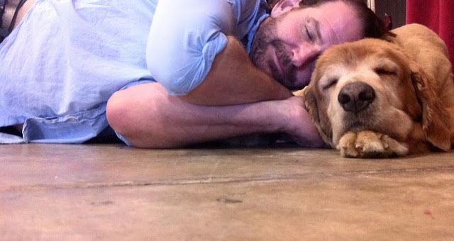 man in blue shirt lying on the floor with his head on a sleeping golden retriever