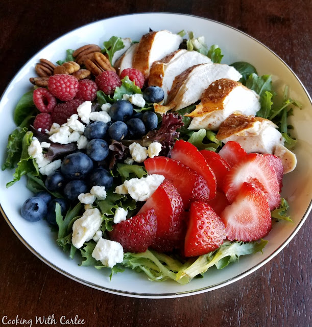 This dinner salad is perfect for Memorial Day, the 4th or July or any day! It is full of berries, cheese, greens, and chicken. There is also a homemade dressing recipe if you'd like! bowl of greens topped with berries, cheese, chicken and nuts