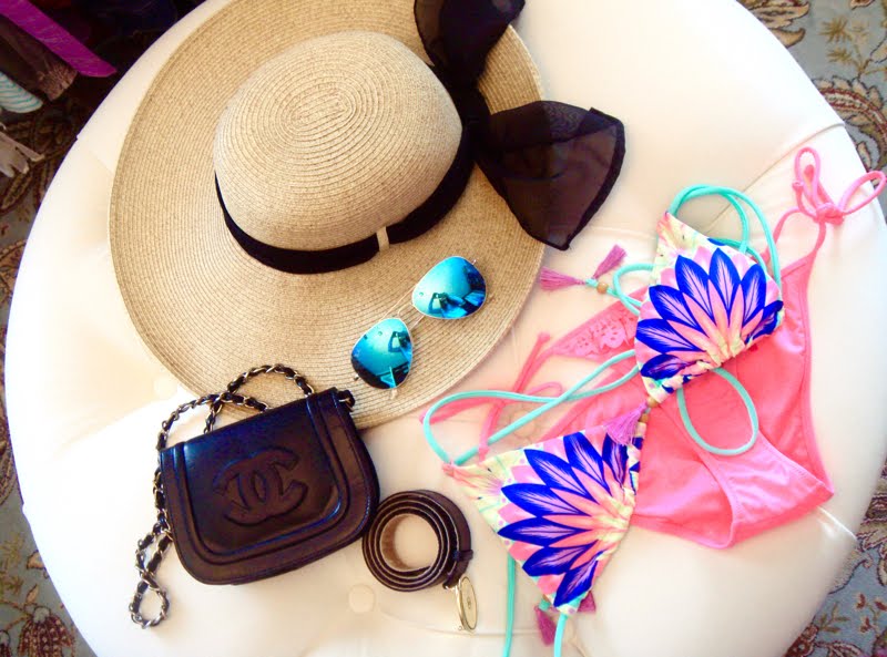 Accessories - hat, ray bans, crossbody bag, belt, and a bathing suit. Accessories laid out on a white ottoman. Straw hat with black bow, blue mirrored sunglasses, black crossbody bag with chain strap, pink and navy palm print bikini and black belt with gold buckel.