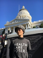Image description: A photograph from the waist-up of a light-skinned person wearing a grey bucket hat and grey shirt that says "Health Care Voter." The person's face is turned to the left as if they are looking into the distance. In the background is the white dome of the California State Capitol Building.