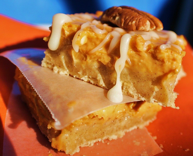 these are an easy how to make pumpkin cheesecake bars. These are creamy filled bars with a cookie crust on the bottom. Drizzled with powdered sugar frosting and pecans on top. They are on orange napkins and a Thanksgiving and Halloween Fall Treat.