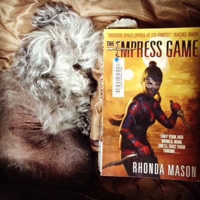 Murchie curls up on a bronze comforter. Beside him is a trade paperback copy of The Empress Game. Its cover features a girl in dark blue and red body paint, her long brown hair drawn back in a ponytail. She wields two curved knives against a golden background lit by two suns. Curled up, Murchie is almost the same size as the book.