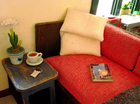 Corner of a miniature sofa, with pillows, a book and a side table holding a cup of tea and a bar of chocolate.