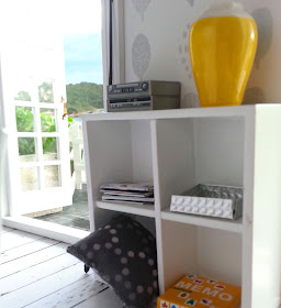 One-twelfth scale modern miniature cube bookcase next to open french doors overlooking a beach. On the shelves is a stereo system, a vase, a pile of magazines, a cushion and a game. Leaning against it is a fishing rod.