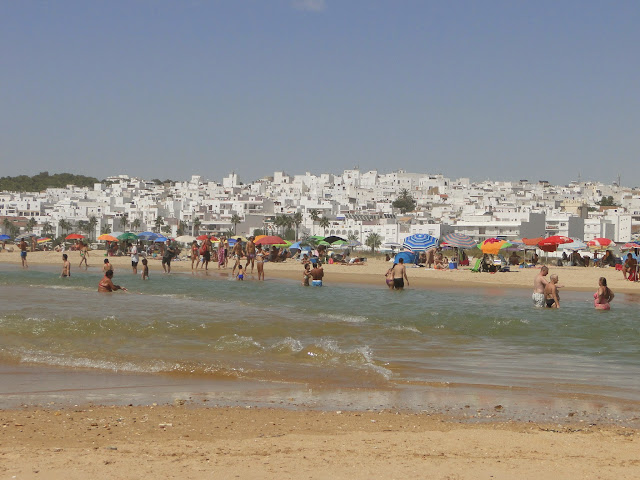La playa y la orilla del mar con oleaje y gente de vacaciones con la ciudad de Conil al fondo.