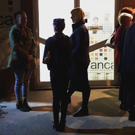 A group of people waiting outside a gallery in the dark. On the gallery door is a sign that reads 'closed'.