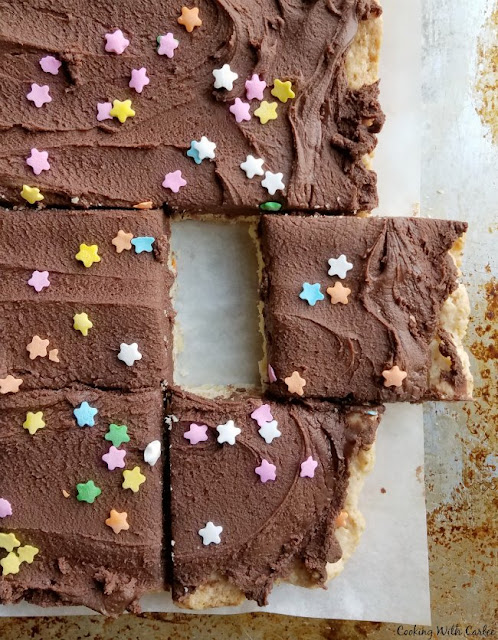 Super soft sugar cookie bars topped with super chocolaty sweetened condensed milk buttercream are a fun, easy and popular treat! Big sheet of sugar cookie bars with chocolate frosting and sprinkles, some bars cut and one slightly pulled away from the rest
