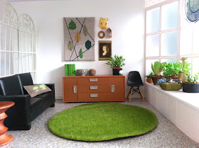 One-twelfth scale modern miniature room with lino floor, white walls and a bay window to the right.There is a black sofa, with an Eames stool next to it, a credenza with art works above it and a black Eames chair to the side of it. The seat of the bay window contains a collection of potted plants.