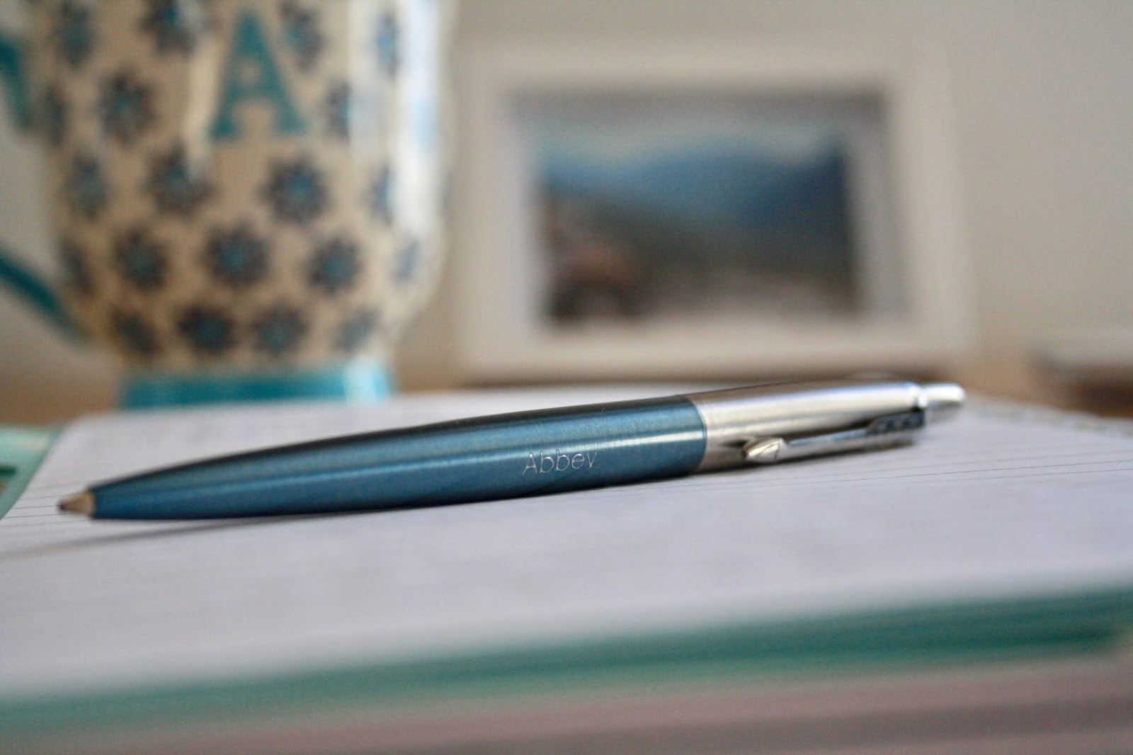 close up of pen resting on open notebook with mug and lamp in background