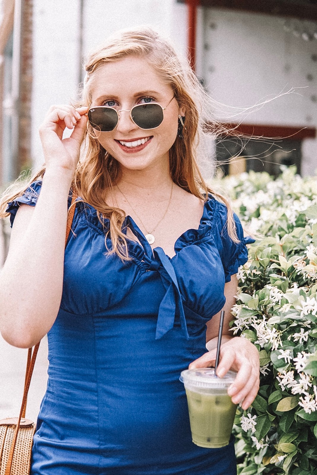 tampa blogger amanda burrows is wearing a navy ruffle dress from francesca's holding a matcha tea from Intermezzo Coffee. She is standing outside Green Bench Brewing. She is wearing Ray Ban Sunglasses and carrying a round rattan circle bag. She is smiling at the camera and standing next to jasmine flowers in St. Petersburg, Florida.