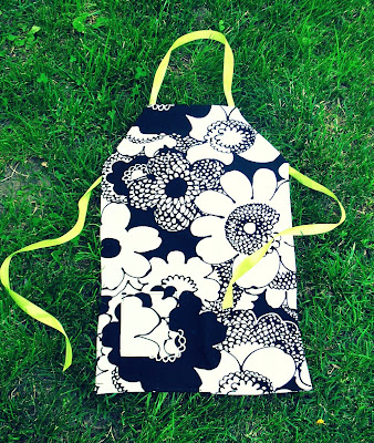 Apron laying on grass with yellow neck hole, and ribbons to tie, the apron has a pocket, and is dark blue and white, and floral patterened
