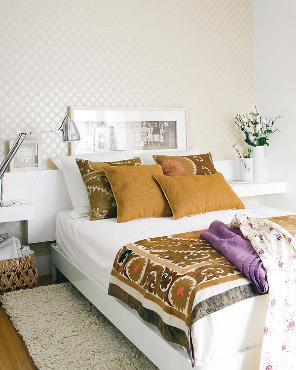 Bedroom in a tiny apartment with one wall covered in wallpaper, wood floor, a shag rug, white floating nightstands on which holds a silver lamp the other a flower arrangement, the bed has sienna colored pillows and a matching patterned throw