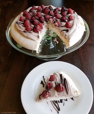 If ever there was a perfect dessert, this might be it. A crisp meringue base that gives way to a marshmallow-y center. Fluffy chocolate whipped cream nestled on top. A smattering of delicious fresh raspberries and a drizzle of chocolate to finish it off. This is a symphony of textures and a delightful mix of flavors. Make one today to find out for yourself! first slice of chocolate raspberry pavlova served with remainder on glass cake plate in background