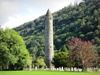 Glendalough Round Tower, Coutny Wicklow, Ireland A tall stone round tower with a cemetery and trees at its feet and a hillside in the background.