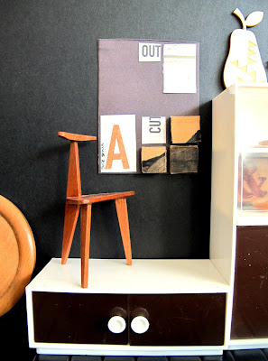 Modern dolls' house miniature collage art work hanging on a black wall. In front of it is an art chair. On the left is a cabinet  with a wooden pear on top of it.