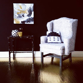 One-twelfth scale modern miniature scene in gold, black, and white comprising a gold-coloured floor and black wall. A grey and black wing chair displays a black and white stitched cushion, and next to it is a black bamboo side table holding three vases in gold, black and white. On the wall above it is a piece of modern art in grey, black, white and gold.