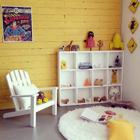 One-twelfth scale modern miniature playroom with concrete floor and yellow brick wall. A white-painted cube bookcase displays toys next to a white chair.
