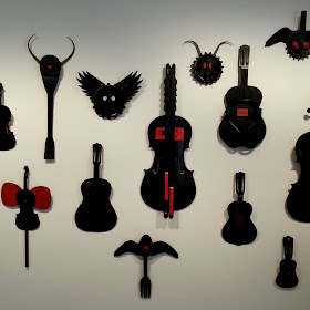 View of an art gallery exhibition with a wall full of Alex Asch assemblage art pieces in black and red, based on various musical instruments.