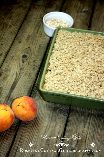 Photo of a green casserole dish on a wooden surface filled with Peach Crisp. A small bowl of oats sits nearby and two ripe, juicy peaches in the foregrouns by RosevineCottageGirls.com