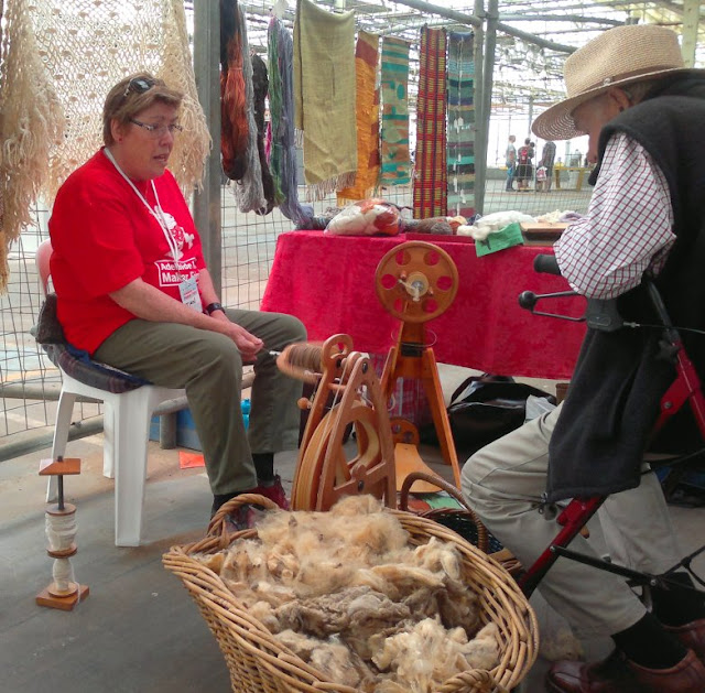 In the foreground is a basket of raw fleece and a spinning wheel behind it.  On the left, Jan is wearing a red t-shirt as she sits spinning while she chats to a gentleman on the right who is sitting on a wheelie walker.