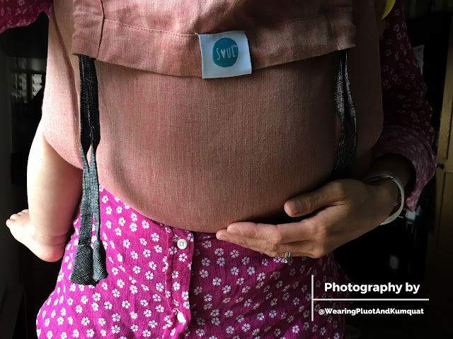 [Image focused on a dusty peach linen onbuhimo snuggling a toddler who is worn on the front of a light tan skin Asian woman who has one hand on the toddler's bum. Bare toddler toes are visible. The disgruntled teething face of the toddler is not visible. The blue SOUL logo on the hood is so cute.]