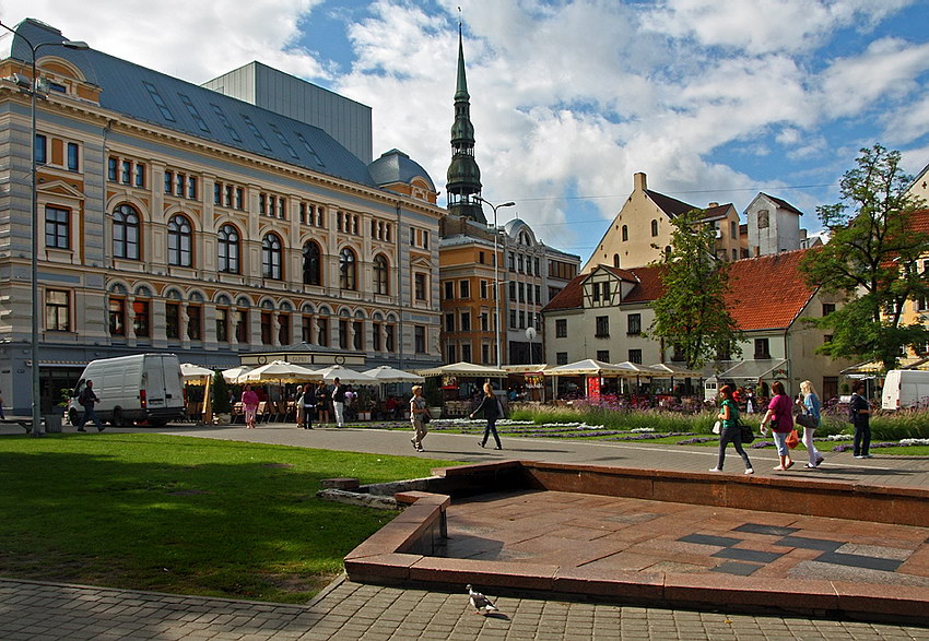 Praça de Livus Vista sobre a praça, com zona relvada e calçada em primeiro plano e ao fundo, toldos e pessoas em circulação. Por detrás, prédios de bela arquitectura e uma torre contra o céu nublado
