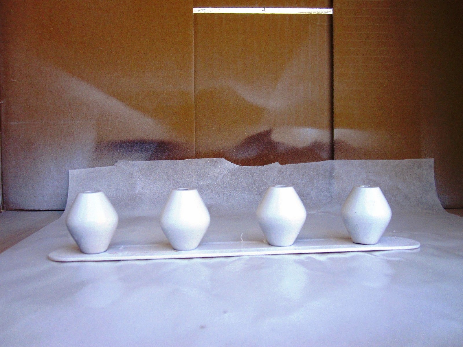 Four geometric wooden beads lined in a cardboard box, having just been spray painted white.