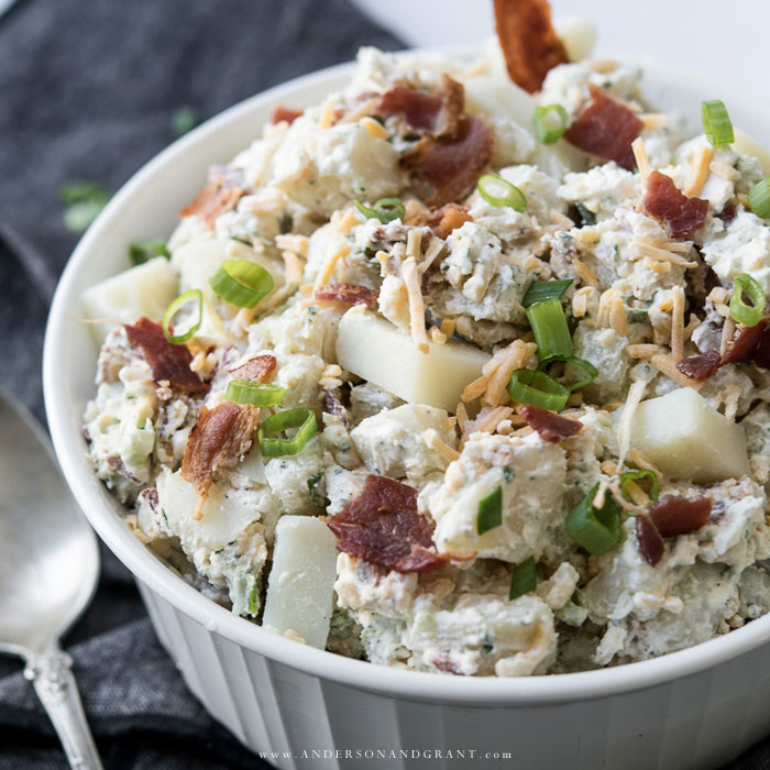 You must try this creamy and flavorful recipe for Potato Salad that includes bacon, ranch seasoning, and sour cream. White dish full of potato salad with bacon, cheese, and onions.