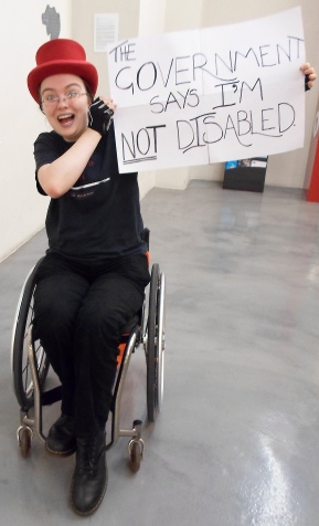 person in wheelchair, dressed all in black except bright red top hat, grinning and holding up a sign that says 'the government says I'm not disabled'