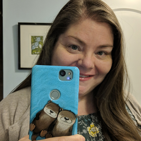 image of my face in a mirror, peeking from behind my phone case, which features cute artwork of two sea otters floating on their backs, holding paws