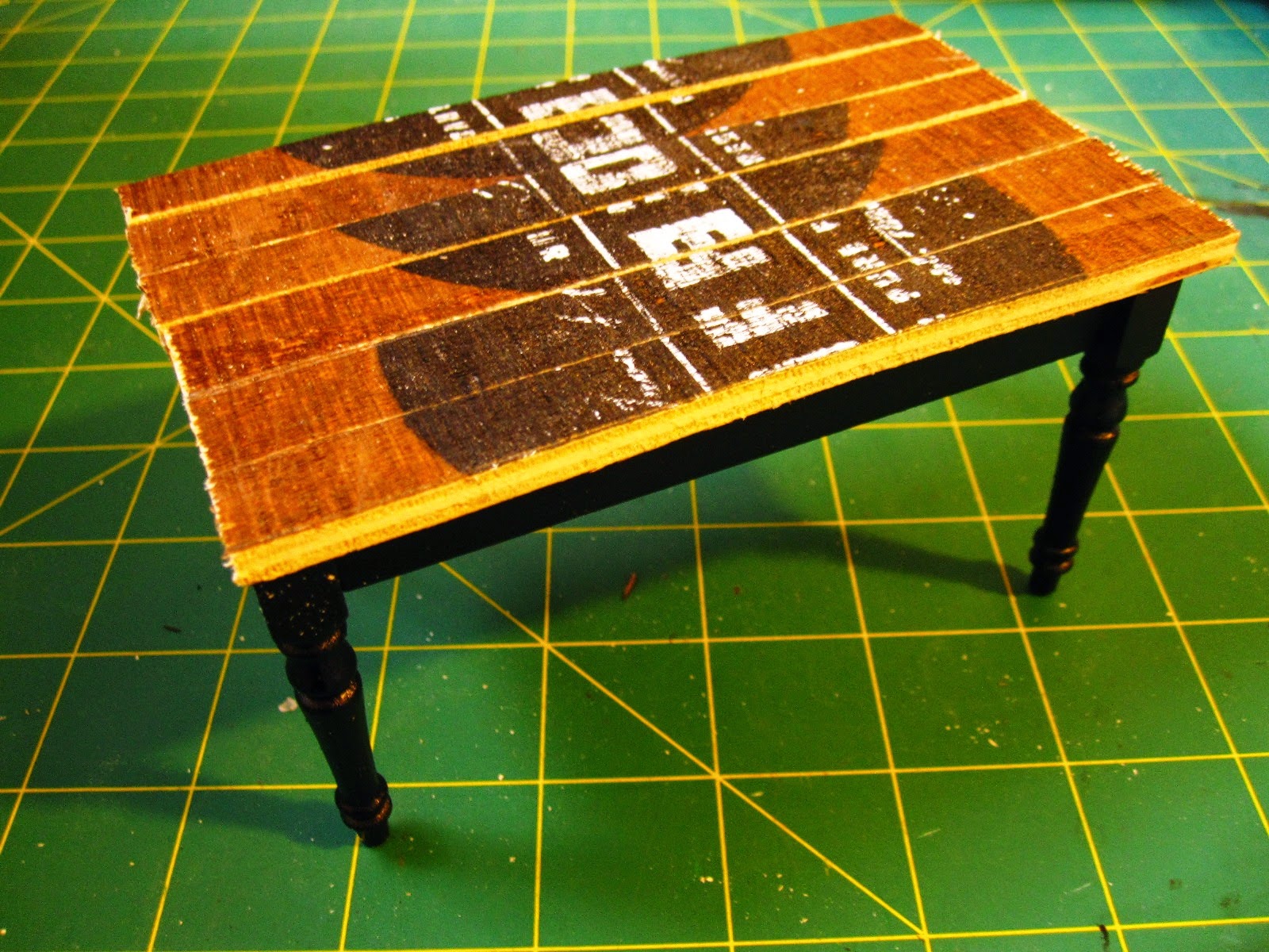 Miniature dining table leg assembly, painted black, topped with a top made of lengths of rustic-looking wood with black printing on them.