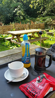 A pot of coffee, some crisps and a bottle of orange drink on an outdoor table.