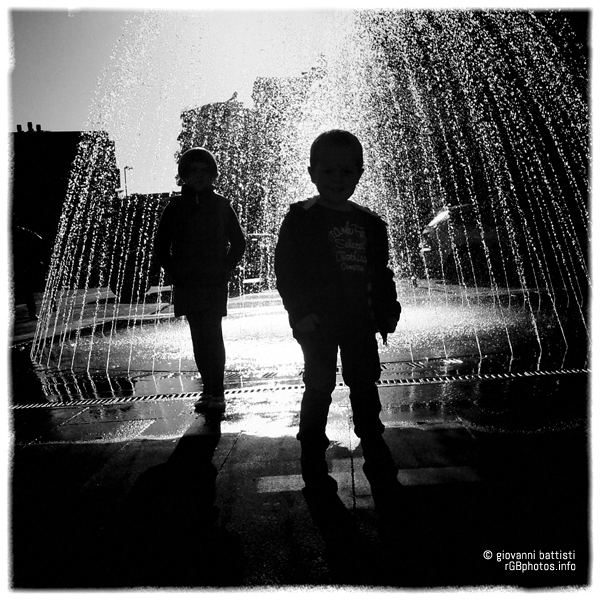 Fotografia controluce di due bambini di fronte ad una fontana cittadina