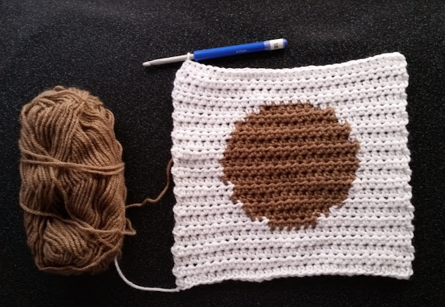 The right side of a white square with a tan circle in the middle. The square is worked in rows using intarsia technique for colour changes. A crochet hook with a blue handle is still inserted into the last stitch of the top row, on the top lef-hand corner of the square. A skein of tan yarn is still joined to the circle and sits to the left of the square.