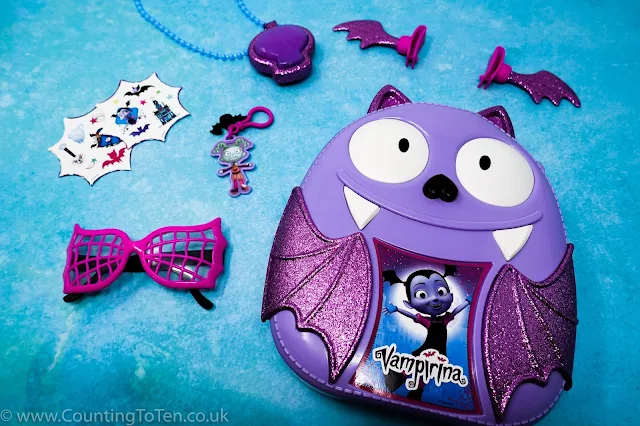 The Vampirina bat shaped backpact surrounded by the items from the set: stickers, sunglasses, necklace, bag clip and hair clips