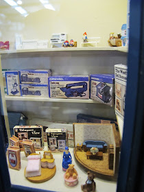 Various dolls' house-related items displayed in the window of a one-twelfth scale toy shop.